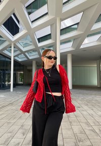 Crvena kardigan s cik-cak uzorkom, crni crop top i široke hlače, postavljeni protiv modernog arhitektonskog pozadine s geometrijskim obilježjima.