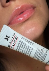 Kiehl's Lip Balm #1 in einer weißen Tube mit schwarzem und rotem Text, gehalten in der Nähe von glänzenden, leicht rosa Lippen auf einem hellen Hautton-Hintergrund.