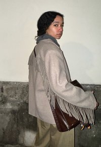 Μπεζ μπουφάν από μαλλί με κρόσσια στους ώμους, συνδυασμένο με γκρι κασκόλ και καφέ δερμάτινη τσάντα. Οι φαρδιές παντελόνες ολοκληρώνουν την εμφάνιση.