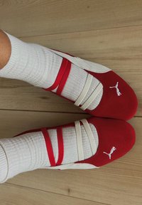 Pantofi sport roșii și albi cu benzi elastice și țesătură texturată, purtați cu șosete albe cu dungi, așezați pe un podea din lemn.