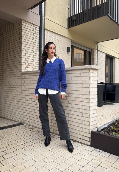 Suéter azul de punto trenzado sobre una camisa blanca con cuello, acompañado de jeans de pierna ancha en gris oscuro y botas de tobillo puntiagudas negras.