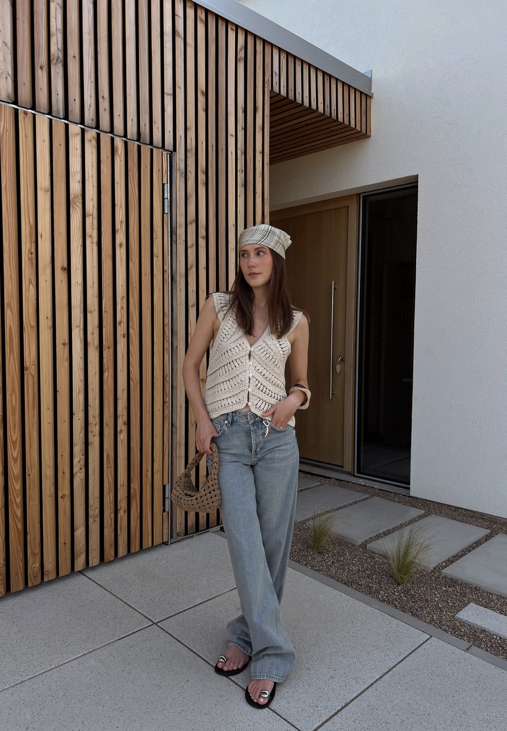 Háčkovaná krémová vesta s výstřihem do V, světle modré kalhoty s širokými nohavicemi, černé sandály se stříbrnými akcenty a vzorovaný šátek, pózující proti dřevěnému obložení.