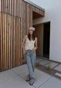 Háčkovaná krémová vesta s výstřihem do V, světle modré kalhoty s širokými nohavicemi, černé sandály se stříbrnými akcenty a vzorovaný šátek, pózující proti dřevěnému obložení.