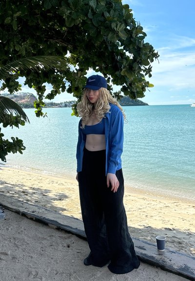 Camisa de mezclilla azul sobre un top corto azul, combinada con pantalones negros holgados. Una gorra de béisbol azul marino completa el atuendo. Playa de arena y agua al fondo.