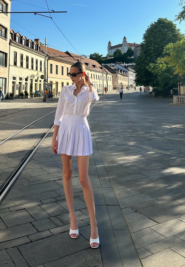 Witte bloes met knopen, gecombineerd met een geplooide witte rok en witte hoge sandalen, tegen een zonnige stadsstraat als achtergrond.