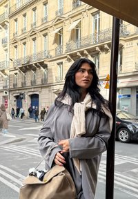 Ženska v sivi plašču in kremastem puloverju z pletenimi vzorci drži bež torbico in stoji ob ulici s klasično arhitekturo.