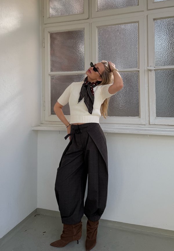 Kvinde iført hvid kortærmet sweater, sorte bukser med vide ben, brune støvler, mønstret tørklæde og solbriller poserer indendørs ved frostede glasvinduer.