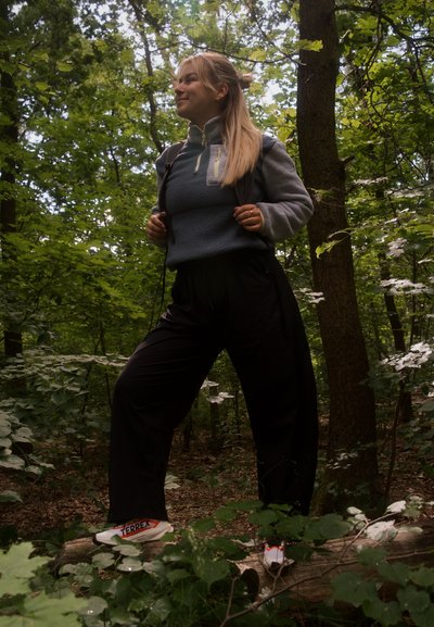 Mujer con mochila de pie sobre un tronco caído en un bosque denso, vistiendo forro polar gris, pantalones negros y zapatillas blancas.