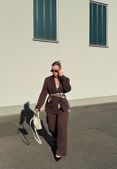 Mujer con traje marrón oscuro, cinturón con patrón y gafas de sol camina por la acera llevando un bolso blanco cerca de una pared blanca con dos ventanas.