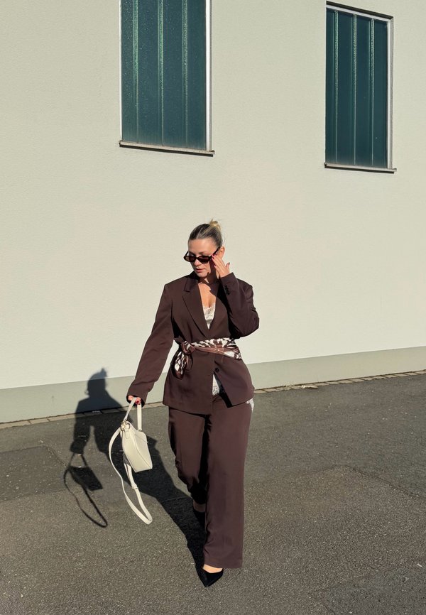 Femme en tailleur marron foncé, ceinture à motifs et lunettes de soleil marche sur le trottoir en portant un sac à main blanc près d'un mur blanc avec deux fenêtres.