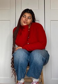 Κόκκινο πλεκτό κασκόλ με κουμπιά, μπλε τζιν σε φαρδιά γραμμή, λευκές δαντελένιες κάλτσες και ροζ παπούτσια. Πλεξούδα μαλλιών και ουδέτερο φόντο.