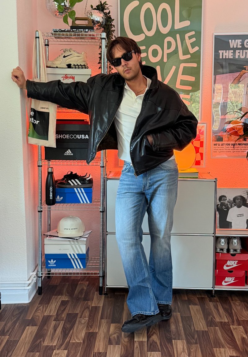 Man wearing sunglasses, black leather jacket, white polo, and blue jeans leaning against a metal shelf with sneakers and posters behind him.