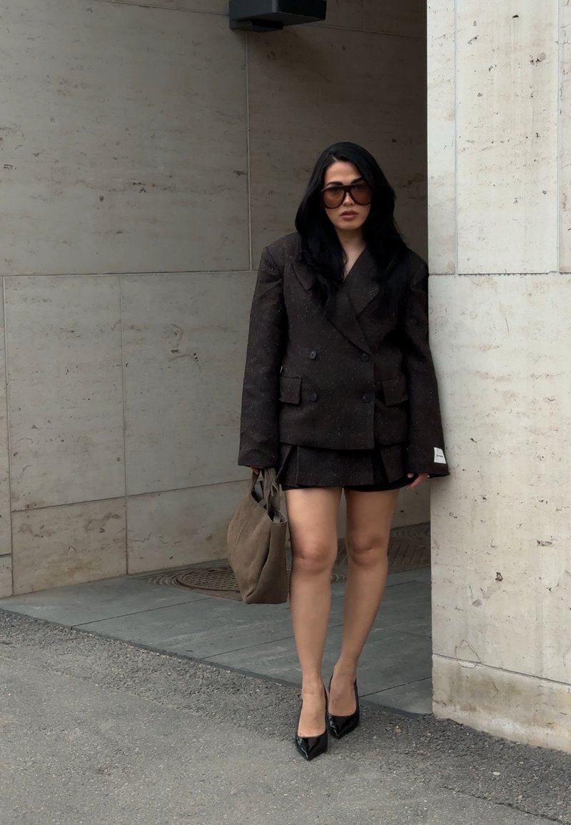 Woman with long dark hair wearing oversized dark blazer and skirt, black heels, large sunglasses, holding beige tote bag, standing by stone wall.
