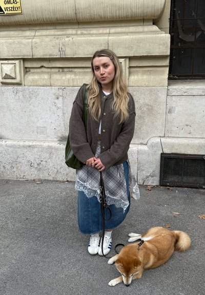 Joven con ropa casual de varias capas de pie junto a una pared de piedra, sujetando la correa de un Shiba Inu color canela que está tumbado en la acera.
