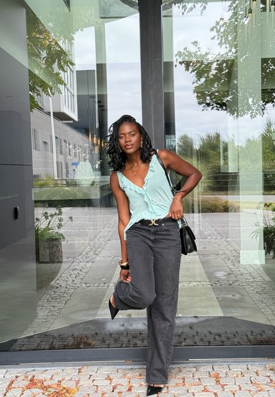 Mujer con pelo trenzado, vistiendo una blusa sin mangas de volantes en azul claro y jeans negros, sosteniendo un bolso negro, ajustando su zapato junto a una puerta de cristal.