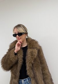 Καφέ ψεύτικο παλτό με ψηλό γιακά, μαύρα ορθογώνια γυαλιά ηλίου και στρώσεις κολιέ, συνδυασμένα με μαύρη μπλούζα και μπλε τζιν.