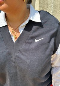 Μαύρος αμάνικος σουτιέν Nike με σχέδιο V-neck, φορεμένος πάνω από ένα λευκό πουκάμισο με γιακά. Ορατές χρυσές στρώσεις κολιέ και σκουλαρίκια.