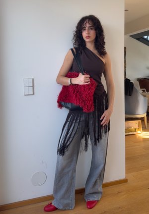 Joven con cabello rizado que lleva un top negro de un solo hombro con flecos, vaqueros anchos grises, zapatos rojos y sostiene un bolso texturizado rojo en el interior.