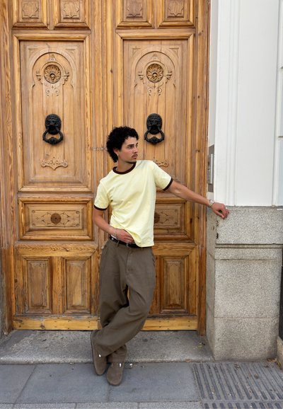Hombre con camisa amarilla y pantalones marrones apoyado contra una pared de piedra con la mano descansando cerca del timbre junto a una gran puerta de madera con aldabas de cabeza de león negras.
