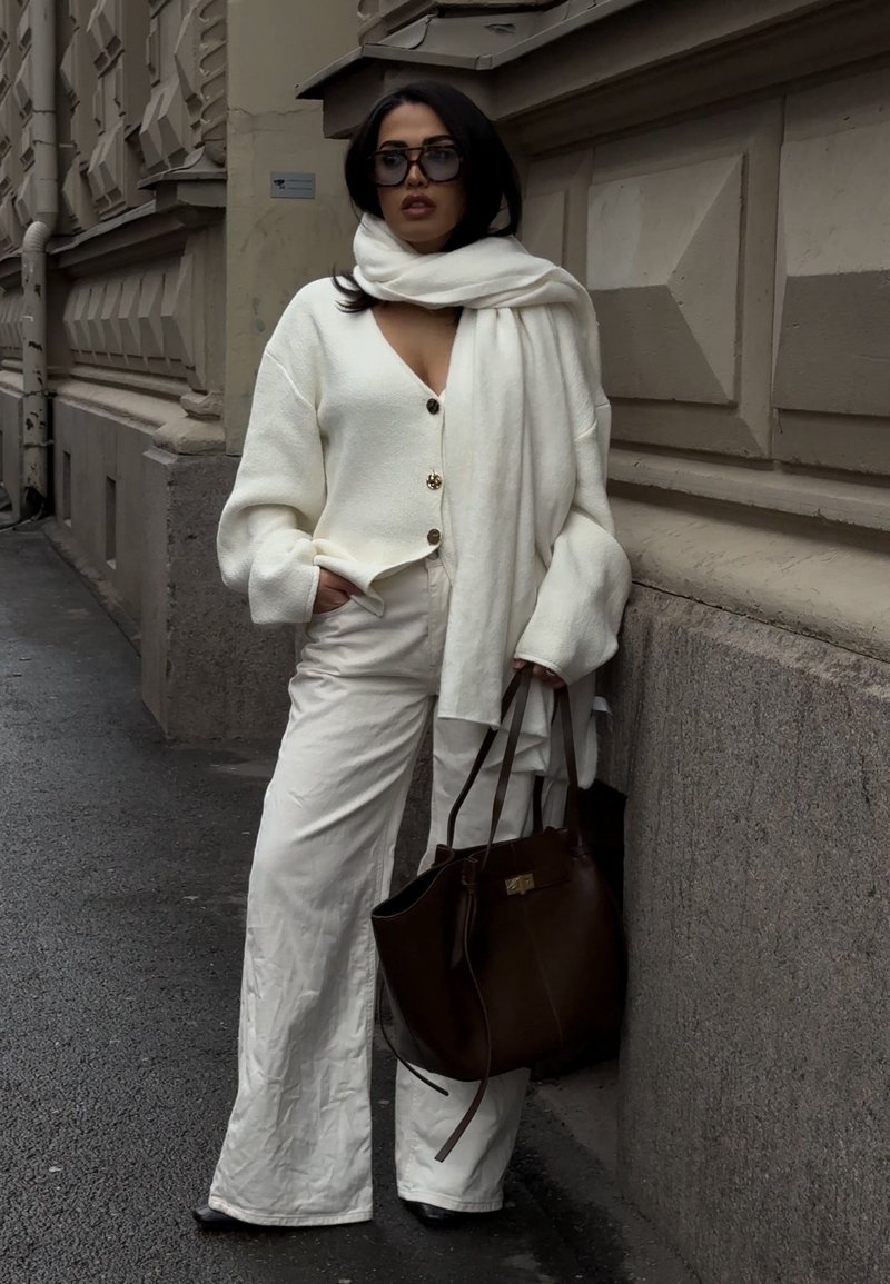 Femme portant un cardigan blanc oversize, un pantalon large assorti et une écharpe, des lunettes de soleil foncées, tenant un grand sac à main marron sur le trottoir de la ville.