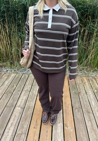 Brūna un balta svītrainā garenpiedurkņu krekls ar apkakli, savienots ar brūnām biksēm un brūniem kurpju sietiem uz koka dēļiem. Klātesoša soma.