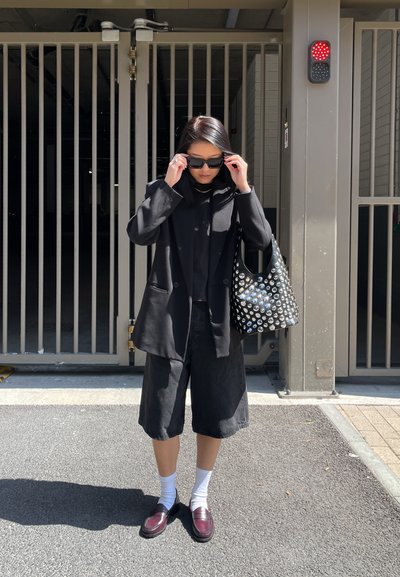 Blazer negro de gran tamaño sobre una blusa y pantalones cortos negros, calcetines blancos, mocasines burdeos y un bolso con tachuelas. Puerta de metal al fondo.