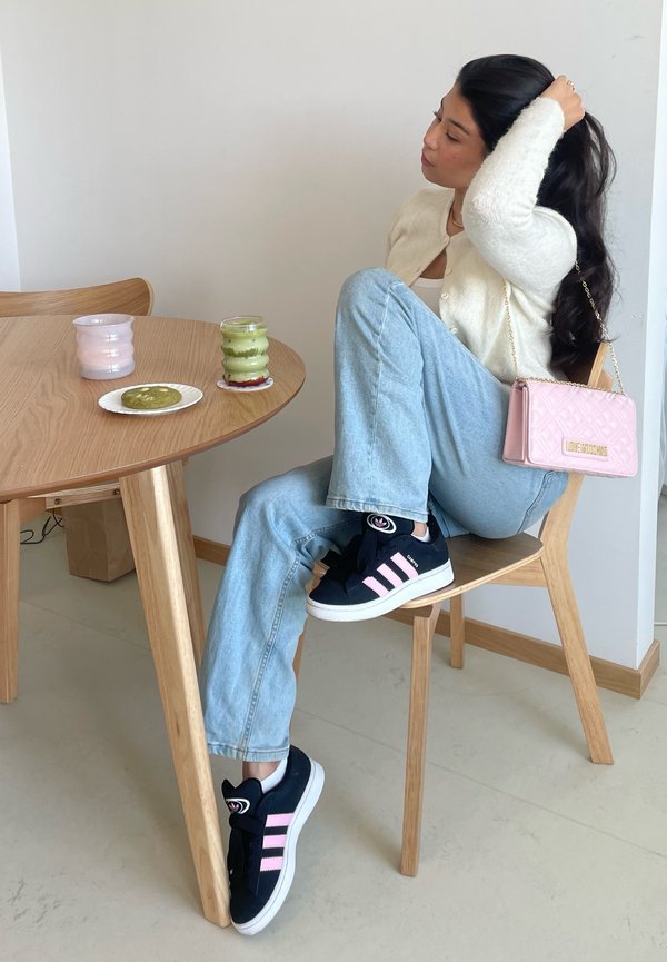 Baskets navy avec des rayures roses, jeans bleu clair et cardigan crème. Un sac à main rose, des verres de boisson et un cookie vert sur la table.