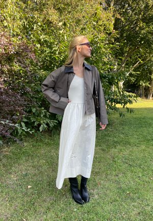 Chaqueta marrón corta con cuello azul, vestido blanco sin mangas y botas negras hasta la rodilla, con un fondo verde y frondoso.