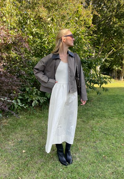 Chaqueta marrón corta con cuello azul, vestido blanco sin mangas y botas negras hasta la rodilla, con un fondo verde y frondoso.