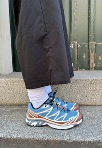 Αθλητικά μπλε και καφέ παπούτσια Salomon τρεξίματος με υφασμένο άνω μέρος, ανακλαστικές λεπτομέρειες και λευκές κορδόνες, φορεμένα με λευκές κάλτσες και μαύρο παντελόνι.