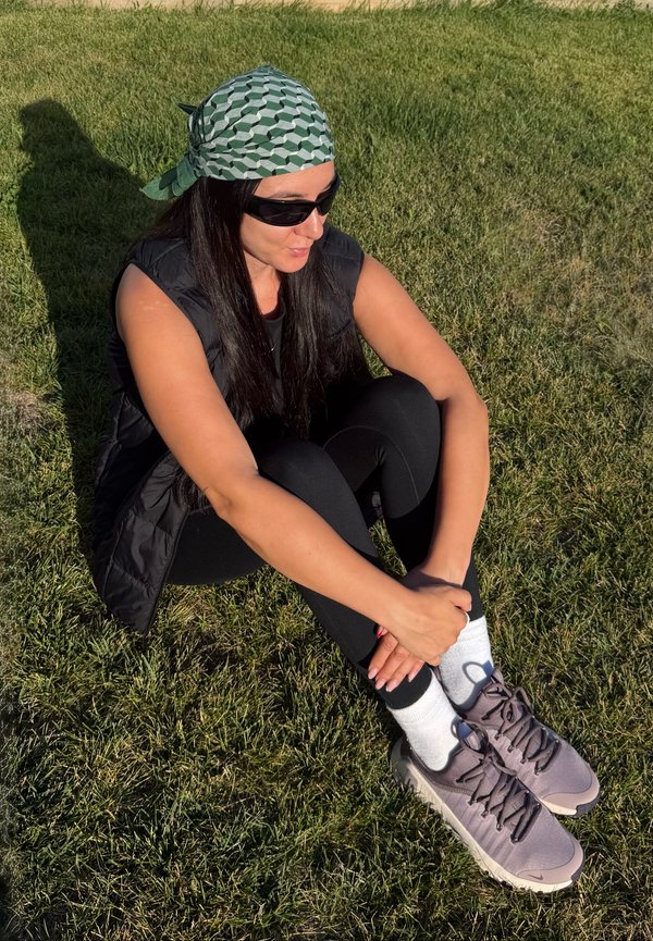 Femme portant des lunettes de soleil, un foulard à motifs, un gilet noir et des leggings, assise en tailleur sur l’herbe sous le soleil, tenant sa cheville.