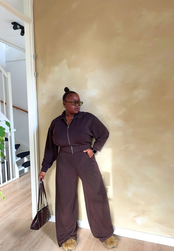 Smeđi trenirka s prugastim uzorkom koja se sastoji od jakne s patent-zatvaračem i širokih hlača, s pahuljastim detaljima na rubovima. Smeđa torba, velike naočale i naušnice.