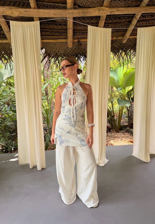 Γυναίκα με λευκό τοπ με μοτίβα τύπου halter και φαρδιά παντελόνα στέκεται σε εσωτερικό χώρο με αιωρούμενες κρεμ κουρτίνες και τροπικά φυτά στο φόντο.