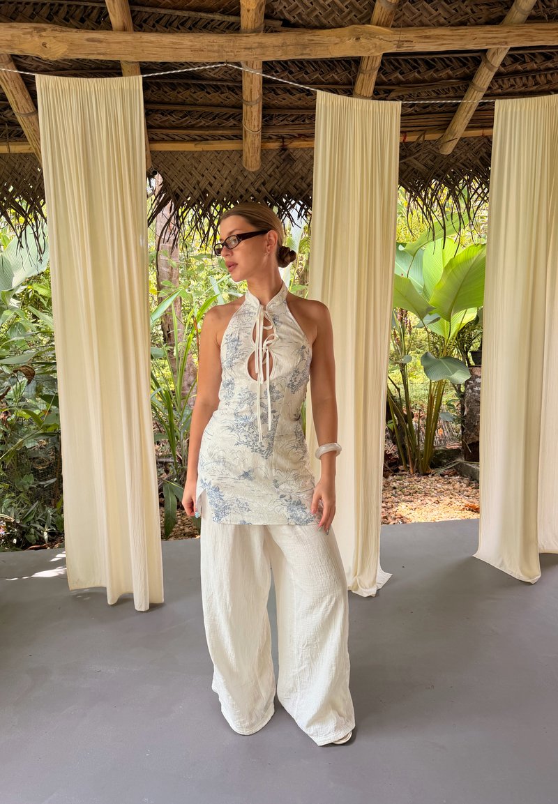 Γυναίκα με λευκό τοπ με μοτίβα τύπου halter και φαρδιά παντελόνα στέκεται σε εσωτερικό χώρο με αιωρούμενες κρεμ κουρτίνες και τροπικά φυτά στο φόντο.