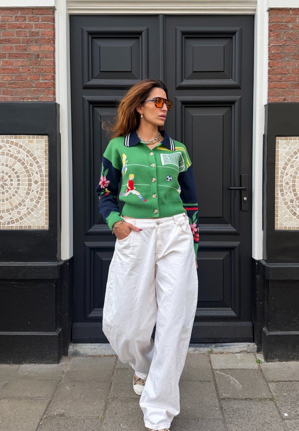Femme portant des lunettes de soleil orange et un pull vert avec des joueurs de football, debout devant une porte noire avec des carreaux de mosaïque sur des murs en briques.