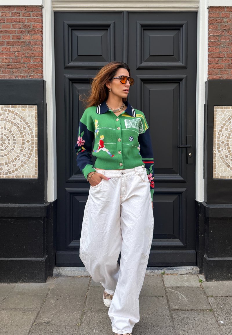 Woman wearing orange sunglasses and a green sweater with soccer players, standing in front of a black door with mosaic tiles on brick walls.
