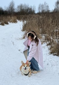 Jeune femme portant un cache-oreilles et un manteau caresse un corgi assis sur un chemin enneigé à côté d'herbes sèches et d'arbres sans feuilles.
