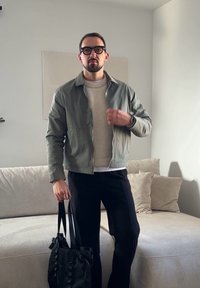 Homme portant des lunettes, une veste grise, un pull beige et un pantalon noir, debout à l'intérieur tenant un sac fourre-tout noir près d'un canapé beige.