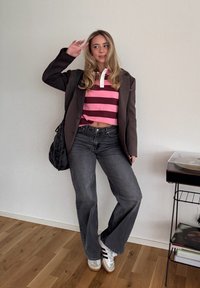 Polo rayé rose et bordeaux, surchemise oversize marron, jeans évasés gris foncé, baskets blanches avec des rayures noires, cheveux ondulés.