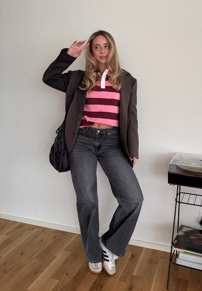 Polo a rayas en rosa y granate, blazer marrón extragrande, vaqueros acampanados gris oscuro, zapatillas blancas con rayas negras, cabello ondulado.