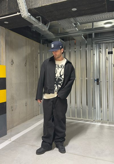 Chaqueta corta negra, suéter gráfico con acentos blancos, pantalones sueltos negros y botas negras. Entorno interior con paredes de concreto y características metálicas.