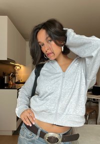 Šedá zipová mikina s mäkkou textúrou, má dlhé rukávy a voľný strih. V kombinácii s vysokými džínsami a veľkou kovovou sponou na opasku.