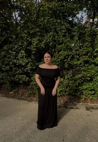 Robe maxi noire, décolleté dénudé, avec un tissu drapé. Texture lisse, corsage ajusté, jupe fluide, portée par une personne devant un fond de verdure.