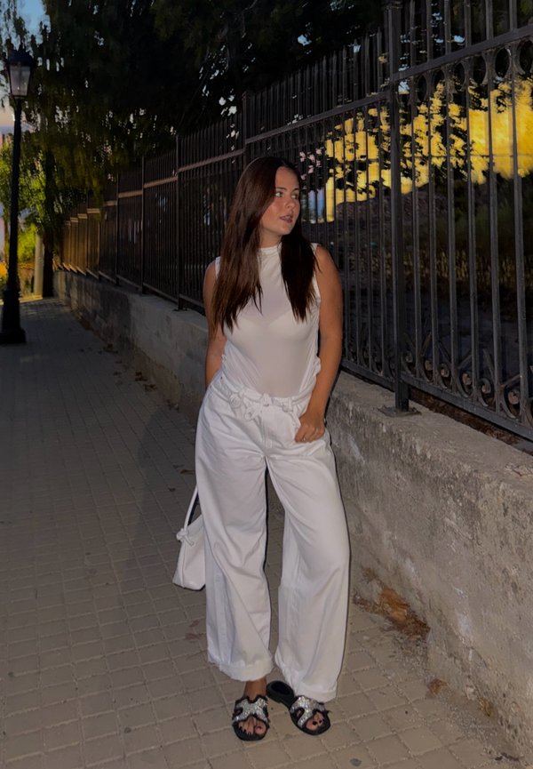 Mujer con top blanco sin mangas y pantalones está en una acera al anochecer, junto a una verja de hierro forjado, mirando hacia un lado con las manos en los bolsillos.