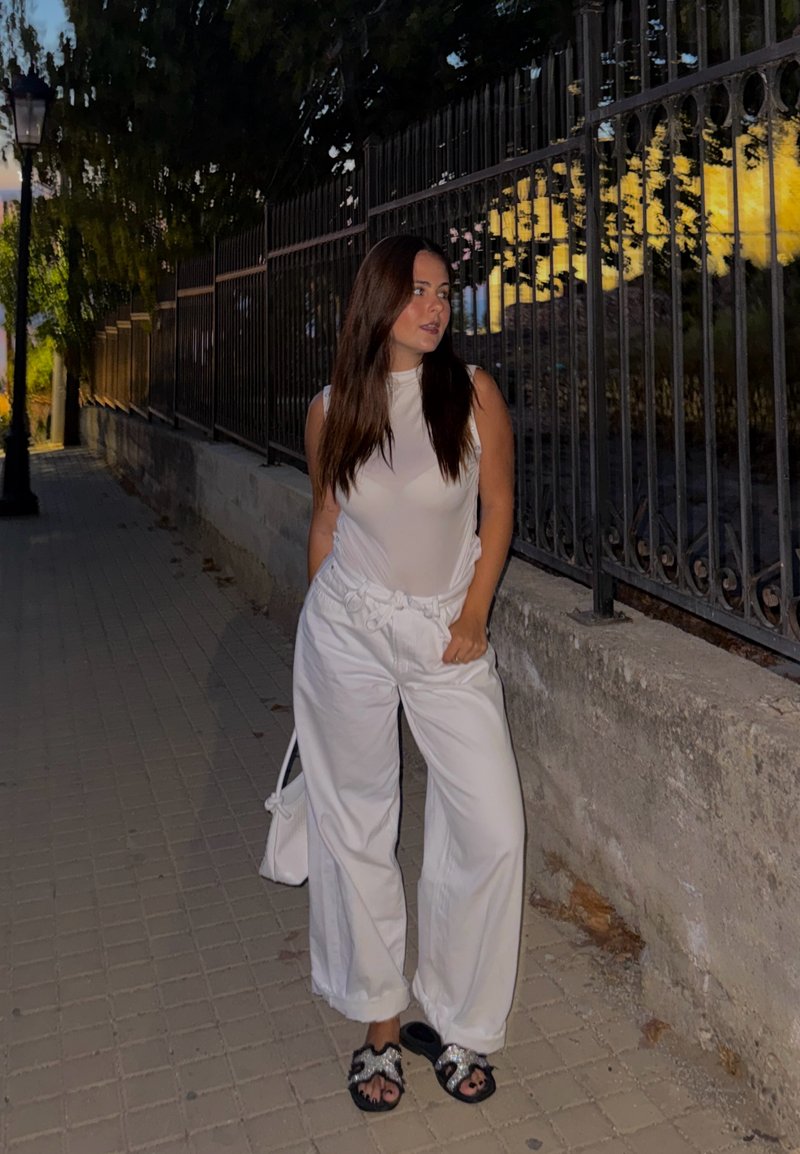 Femme en haut blanc sans manches et pantalon se tient sur un trottoir au crépuscule, à côté d'une clôture en fer forgé, regardant sur le côté, les mains dans les poches.