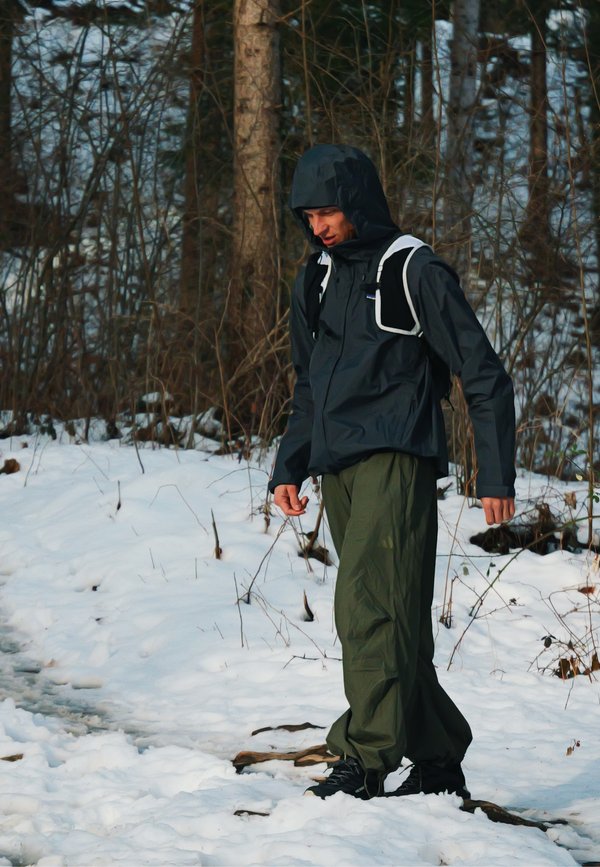 Hombre con chaqueta negra con capucha y pantalones verdes caminando por un sendero nevado en el bosque, con árboles desnudos al fondo.