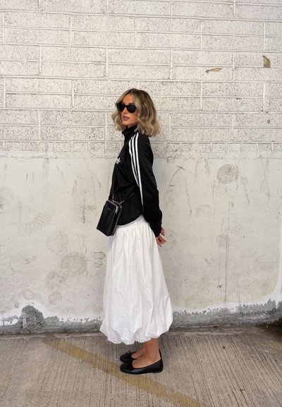 Chaqueta negra con cremallera y rayas blancas, falda blanca hasta la rodilla, zapatos planos negros y gafas de sol; de pie contra una pared blanca texturizada.