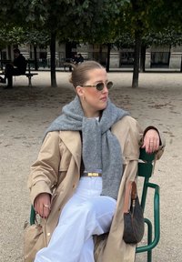 Beiger Trenchcoat über einem grauen Pullover, weißen weit geschnittenen Hosen und Sonnenbrille. Hält eine braune Handtasche mit dunklem Muster.