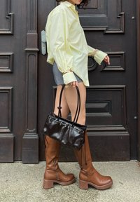 Camisa amarela oversized com mangas longas, calções cinzentos, botas castanhas até ao joelho e uma pequena bolsa preta com duas alças, posicionada junto a uma porta de madeira.