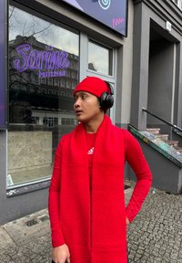 Gorro vermelho, cachecol longo e camiseta de manga longa justa feita de um material suave. Usando auscultadores em cima da orelha, de pé à frente de uma pastelaria.